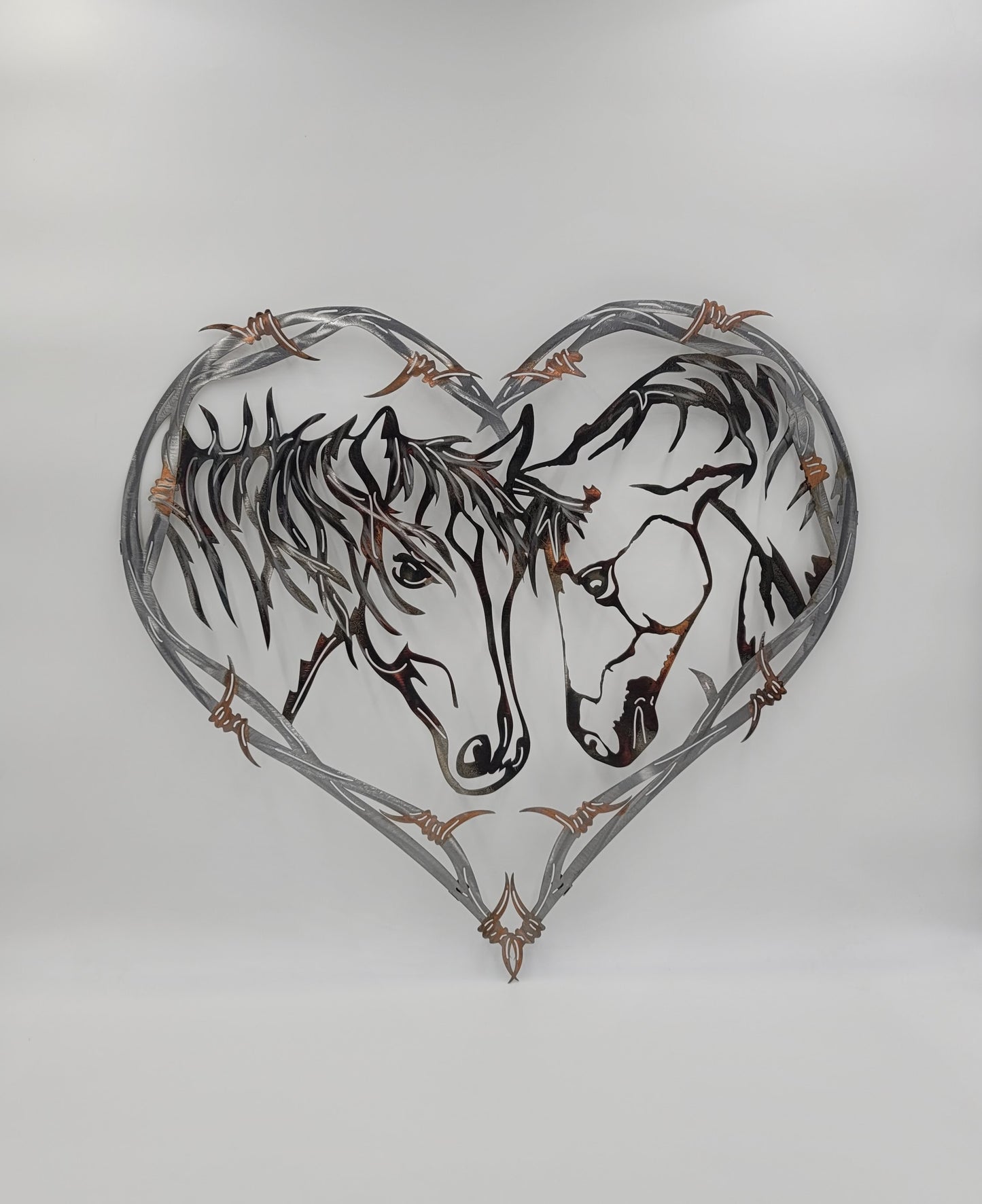 Two Horses in a Barbwire Heart Wall Hanging. It's made from 100% American made cold rolled 18-gauge sheet metal. Measures 24 1/2 Inches High x 25 1/2 Inches Wide x 18 Gauge. Brackets on back for hanging. Four Color Options Available; Colored-Rustic, Black, Hammer Tone, Bare Metal.