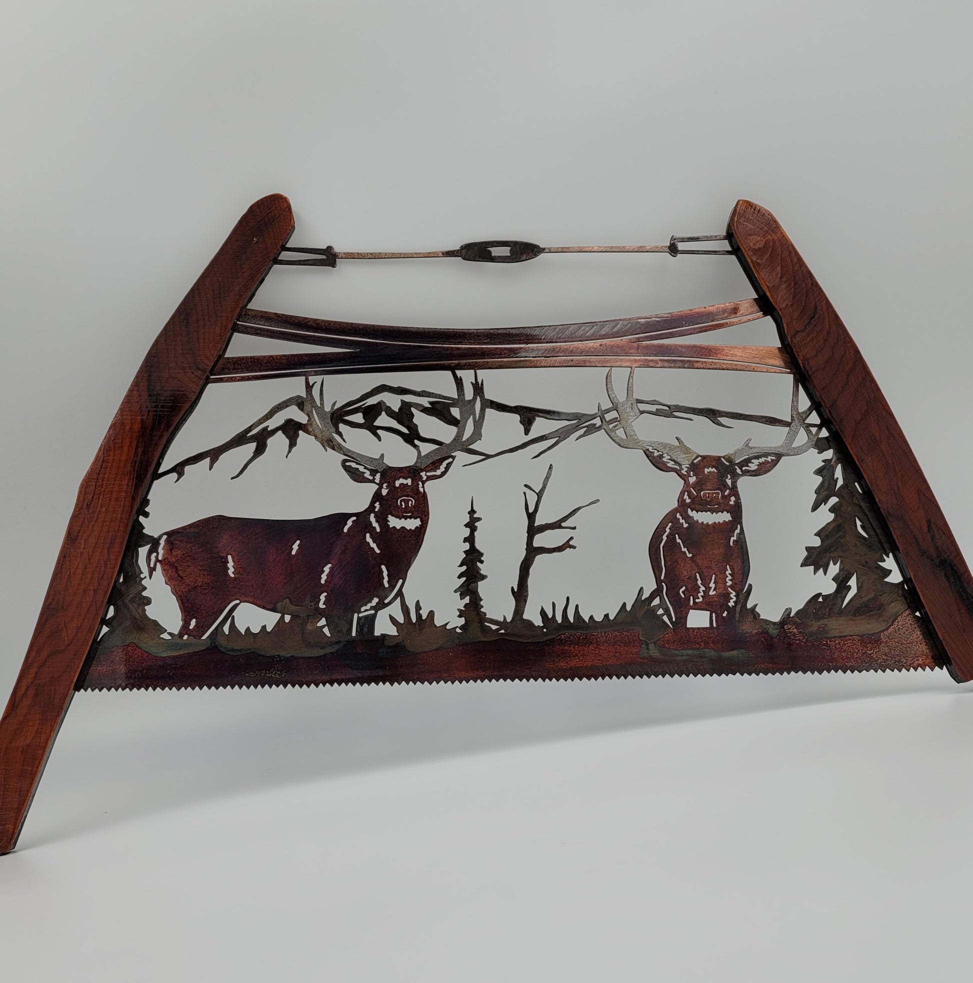A decorative metal bow saw wall hanging featuring two deer in a forest setting, with a wooden handle.