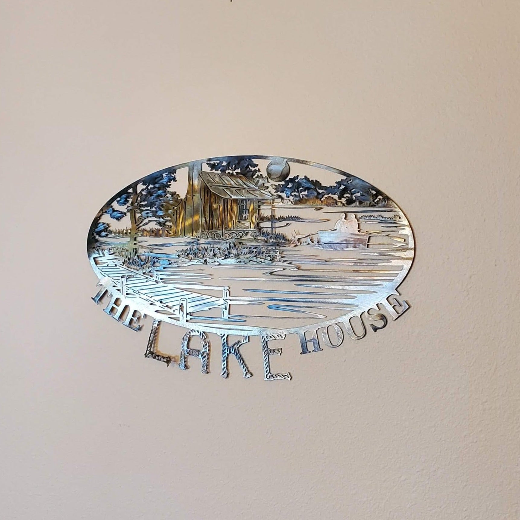 The Lake House Sign features a rustic cabin nestled next to a river. There's a dock on the water, trees, mountains, and a moon in the background. On the bottom of the sign it says The Lake House. Measures 20 inches wide by 15.5 inches high x 18 gauge. All our signs are sprayed with a thick layer of Varathane.