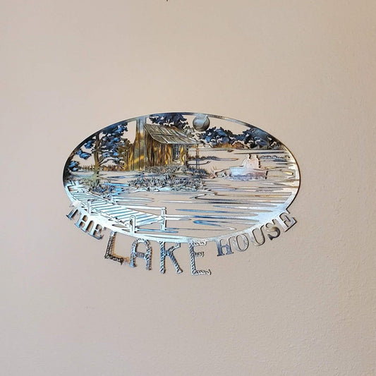 The Lake House Sign features a rustic cabin nestled next to a river. There's a dock on the water, trees, mountains, and a moon in the background. On the bottom of the sign it says The Lake House. Measures 20 inches wide by 15.5 inches high x 18 gauge. All our signs are sprayed with a thick layer of Varathane.