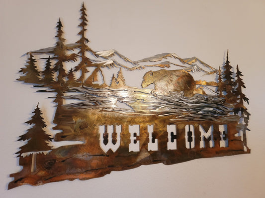 Bear Welcome Sign features a bear walking through a river with mountains and trees in the background. The word Welcome is on the bottom. Made from 18-gauge cold rolled sheet metal. Available in four colors. Colored which is very rustic looking. Black, hammer tone, and bare metal. Measures 17 1/4 inches high by 26 1/2 inches long. Sprayed with several layers of varathane. Hangs via nails not included. Can be hung on porch as long as it's kept away from rain and snow. Gift wrapping available at checkout.